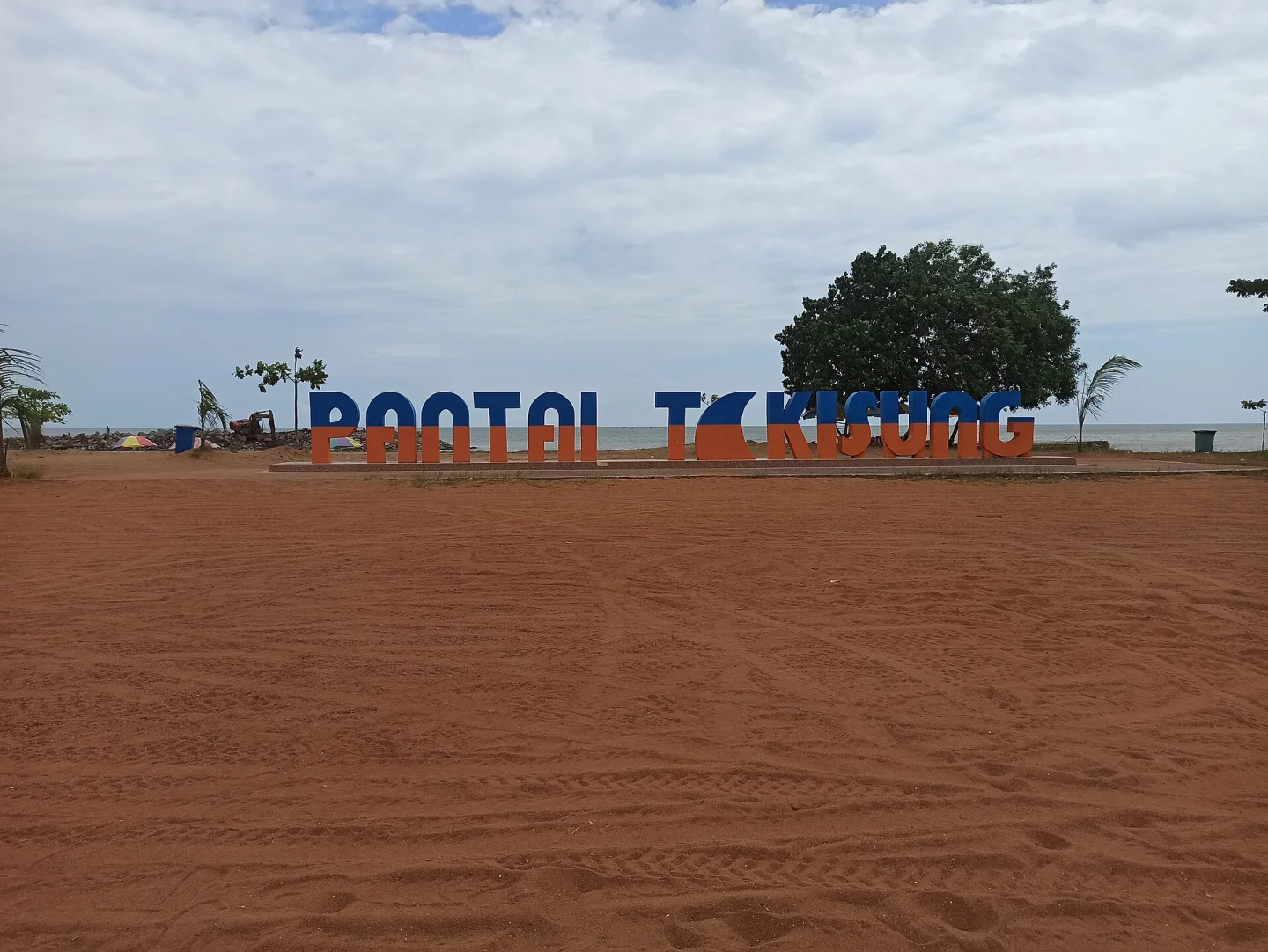 Tulisan besar PANTAI TAKISUNG berwarna biru dan oranye di hamparan pasir, dengan laut dan pohon rindang di belakangnya