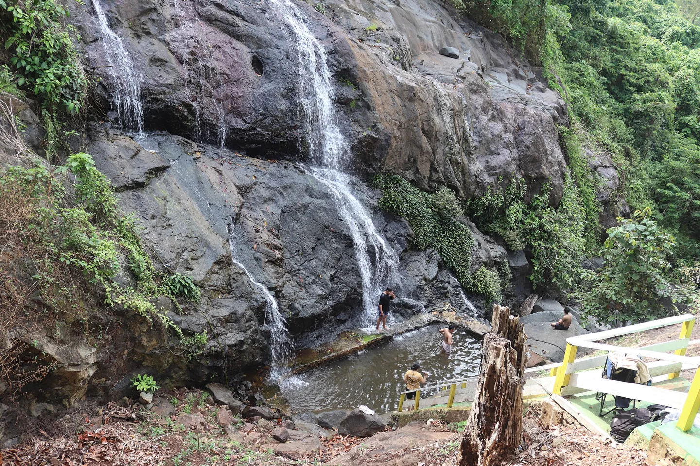 Air terjun mengalir di tebing batu berlumut ke kolam kecil tempat beberapa pengunjung berendam, dengan tangga berpagar kuning di sisi kanan