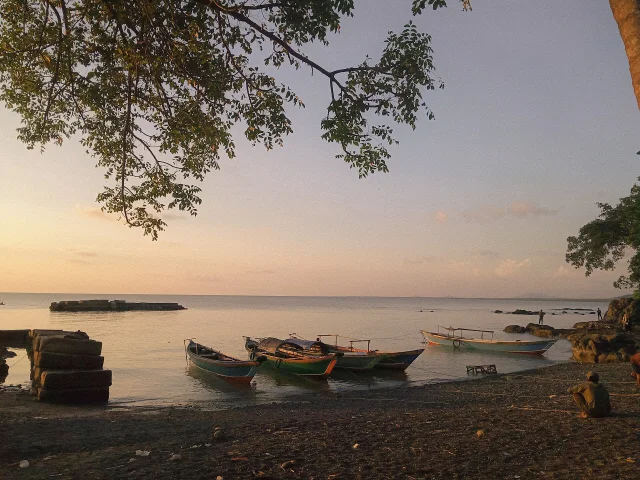 Perahu-perahu nelayan berjajar di tepi Pantai Tanjung Dewa yang berbatu saat matahari rendah, dinaungi dahan pohon